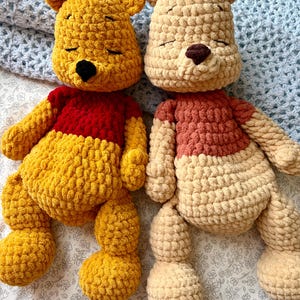 May include: Two handmade crochet teddy bears. One bear is yellow with a red shirt and black nose. The other bear is beige with a pink shirt and brown nose. Both bears have closed eyes and are lying on a floral patterned surface.
