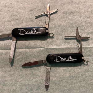 May include: Two black multi-tools with silver blades and scissors. The tools have the name "Daniel's" in white script. One tool has a blade and a file extended. The other tool has a blade and a file extended, and the scissors are open.