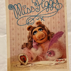 May include: A rectangular print featuring Miss Piggy with long brown hair, wearing a pink feathered garment and purple gloves, holding a purple quill pen. The text Miss Piggy is written in blue script above, with a heart-shaped frame containing Kermit.
