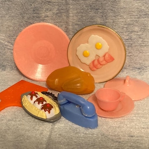 May include: A collection of vintage toy food and kitchen items in various colors. Includes a pink plate, a plate with eggs and bacon, a toy turkey, a banana split, a blue iron, a cup and saucer, and a lid.