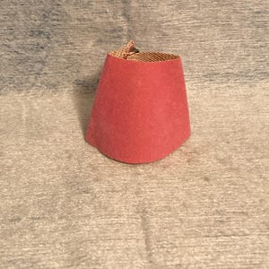 May include: A small, red fez hat with a rounded top and a flat, circular base. The interior lining is a woven, tan fabric. The hat is made of a soft, felt-like material.