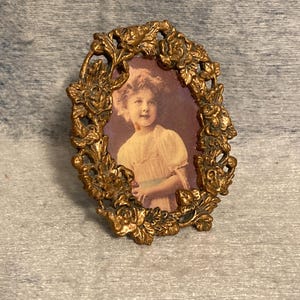 May include: An oval-shaped picture frame with a metallic gold finish and a detailed relief pattern of roses and leaves. The frame contains a portrait of a young person with light-colored hair wearing a light-colored top with short sleeves.