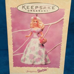 May include: A Keepsake Ornament box featuring a Springtime Barbie doll. The doll wears a white dress with pink floral patterns and carries a basket. The box has a pink background with the words "Springtime Barbie" and "New Collector's Series".