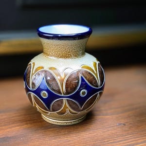 May include: A small, decorative ceramic vase with a rounded body and a narrow neck. The vase features a brown and tan speckled background with a blue and brown floral pattern. The rim is blue, and the vase sits on a wooden surface.