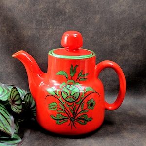 Könnte beinhalten: Eine rote Keramik-Teekanne mit grünem Blumendesign. Die Teekanne hat einen passenden Deckel mit kleinem Knopf und grünem Rand. Griff und Ausguss sind gebogen. Das florale Design umfasst eine große Blume und Blätter.