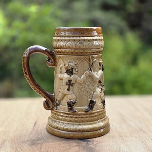Könnte beinhalten: Ein beigefarbener Bierkrug aus Keramik mit einem braunen Henkel. Der Krug hat ein detailliertes Relief von Figuren in historischer Kleidung mit einem Kreuzemblem auf ihren Tuniken. Henkel und Rand haben eine dunklere braune Glasur.
