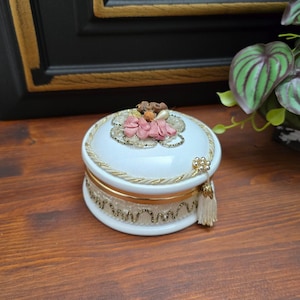 May include: A white ceramic trinket box with a gold trim and a pink and white floral design on the lid. The box has a tassel.