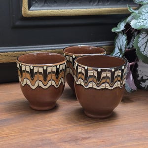 Könnte beinhalten: Drei braune Keramikbecher mit einem geometrischen Muster in Schwarz, Weiß und Braun. Die Becher haben eine abgerundete Form und eine leicht ausgestellte Lippe.