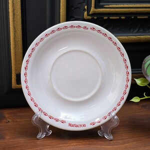 May include: A white saucer with a red decorative border and the word "Mariacron" printed in red. The saucer is displayed on a clear plastic stand. The design features a repeating pattern of stylized shapes.