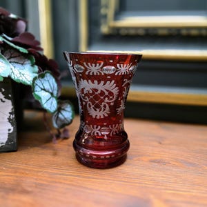Könnte beinhalten: Eine rubinrote Glasvase mit aufwendigen weißen geätzten floralen und geometrischen Mustern. Die Vase hat einen ausgestellten oberen Rand und einen abgerundeten Boden. Die Vase ist etwa 13 cm hoch und steht auf einer Holzoberfläche.