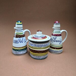 May include: A set of three ceramic kitchen containers. Two bottles with handles and the word "Vel" and "Essig" in script, and a small jar with a lid. The containers are white with colourful stripes and patterns.