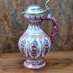 Könnte beinhalten: Dekorative Keramikkrug mit silbernem Deckel und Griff. Der Krug hat eine weiße Basis mit rosa, blauen und grünen Blumenmustern. Der Griff ist ein verdrehtes rosa Design. Der Krug wird wahrscheinlich zum Servieren von Getränken verwendet.