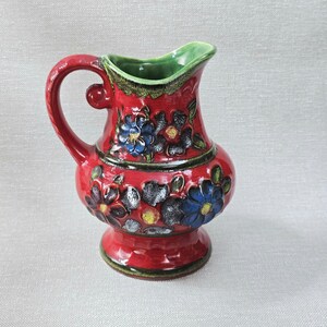 May include: A red ceramic pitcher with a handle and a flared spout. The pitcher is decorated with blue and black floral designs and has a green interior. The pitcher is approximately 7 inches tall.