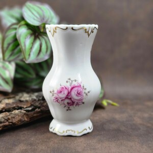 May include: A small, white porcelain vase with a flared top and a pedestal base. The vase features a floral design of pink roses and gold trim. The vase is approximately 5 inches tall.
