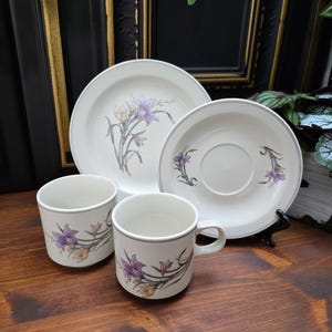 May include: A white ceramic tableware set with a floral design in purple and yellow. The set includes two mugs, a saucer, and a dinner plate. Each item has a grey trim.