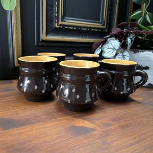 Könnte beinhalten: Satz von sechs braunen Keramikbechern mit weißem Tupfenmuster und gelbem Rand. Die Becher sind in einem Halbkreis auf einer Holzoberfläche angeordnet.