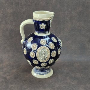 May include: A dark blue ceramic jug with a cream-coloured handle, spout, and base. The jug is decorated with cream-coloured floral and circular patterns, and a central emblem. The jug is approximately 30 cm tall.