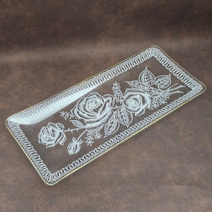 May include: Rectangular glass serving tray with a white rose design. The tray has a gold trim and a decorative white border. The rose design includes three roses, leaves, and stems. The tray is clear and sits on a brown surface.