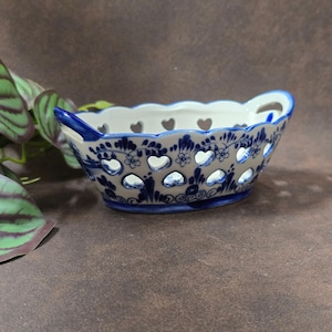 Könnte beinhalten: Ein blau-weißer Keramikkorb mit herzförmigen Aussparungen und Griffen. Der Korb hat einen gewellten Rand und florale und wirbelnde Designs. Der Innenraum ist weiß, und der Außenraum ist blau und weiß.