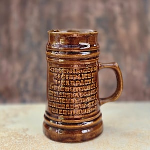 Könnte beinhalten: Ein brauner Bierkrug aus Keramik mit Henkel und geprägtem Text. Der Krug hat eine zylindrische Form mit breiterem Boden und schmalerem Oberteil. Der Text scheint auf Polnisch zu sein. Der Krug ist dunkelbraun.