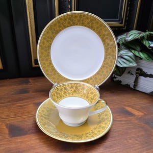 May include: A set of gold and white teacups and saucers with a delicate floral pattern. The set includes a teacup, saucer, and a larger plate.