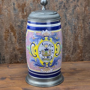 May include: A decorative ceramic beer stein with a metal lid and base. The stein features a floral design in yellow, blue, and pink. The background is a dark brown color.