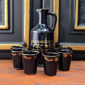 Könnte beinhalten: Ein dunkelblauer Keramikkrug mit Henkel und sechs passenden Schnapsgläsern. Krug und Gläser haben goldfarbene Zierbänder und Muster. Das Set wird auf einer Holzoberfläche präsentiert.