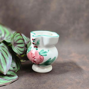 Könnte beinhalten: Kleine, weiße Keramikvase mit floralem Design. Die Vase hat eine rosa Rose und grüne Blätter mit einem türkisfarbenen Rand. Die Vase ist ein dekorativer Gegenstand, wahrscheinlich für kleine Blumen oder zur Präsentation.