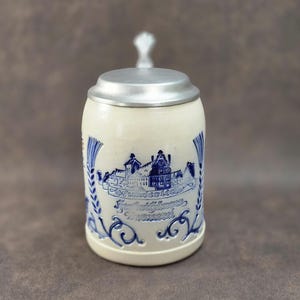 May include: Cream-colored ceramic beer stein with a silver lid and handle. The stein features a blue illustration of a town and text. Decorative blue wheat stalks and scrollwork adorn the sides. A classic German-style beer mug.