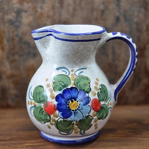 Könnte beinhalten: Ein Keramikkrug mit gesprenkelter cremeweißer Glasur, verziert mit einer handgemalten blauen Blume in der Mitte, umgeben von grünen Blättern und kleinen roten Akzenten. Der Rand und der Henkel haben blaue Verzierungen und der Boden einen blauen Ring.