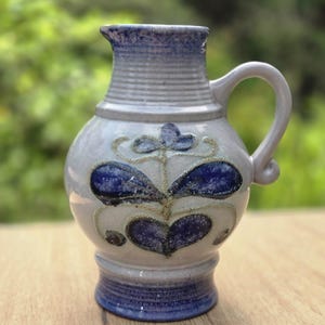 Könnte beinhalten: Ein Keramikkrug mit hellgrauem Grundkörper und blau glasiertem Rand sowie Fuß. Der Körper zeigt ein handgemaltes florales Motiv in Dunkelblau mit strukturierten goldfarbenen Akzenten. Er verfügt über einen geschwungenen Henkel und einen Ausguss.
