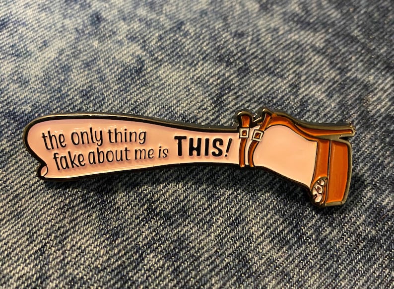May include: A gold-colored enamel pin with a pink leg and a brown high-heeled shoe. The leg has the text "the only thing fake about me is THIS!" written on it.