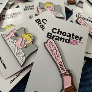 May include: Two enamel pins featuring a woman with a crown and a pair of legs in high heels. The pins are white with pink and black accents. The text on the pins reads "Cheater Brand" and "The only thing fake about me is THIS!"