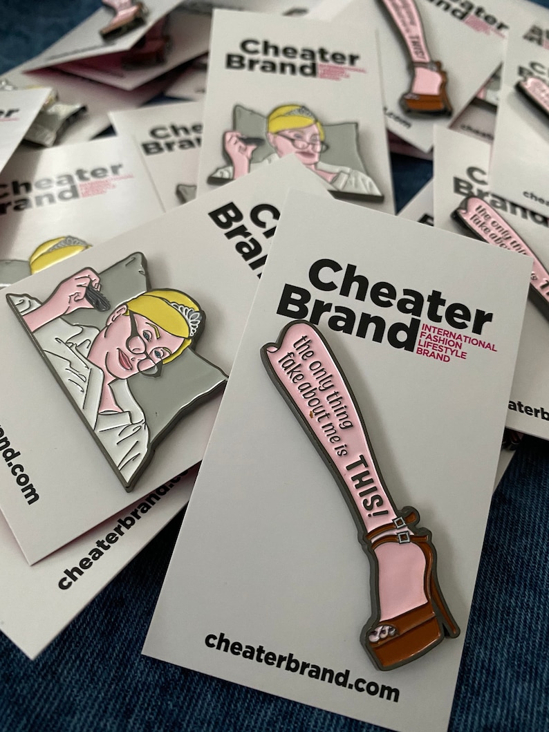 May include: Two enamel pins featuring a woman with a crown and a pair of legs in high heels. The pins are white with pink and black accents. The text on the pins reads "Cheater Brand" and "The only thing fake about me is THIS!"