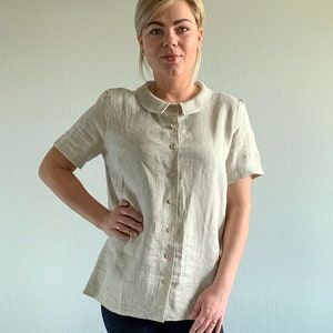 May include: A woman wearing a light beige linen shirt with a peter pan collar and short sleeves. The shirt has a button-up front and is tucked into dark blue jeans. She is wearing white sneakers with gold accents.