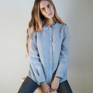 Op de afbeelding: Een blauw-wit gestreept overhemd met een ronde kraag. Het shirt met lange mouwen heeft een verticaal streepjespatroon en is aan de voorkant dichtgeknoopt. Het shirt wordt gecombineerd met een grijze jeans. Het model zit op een houten kruk.