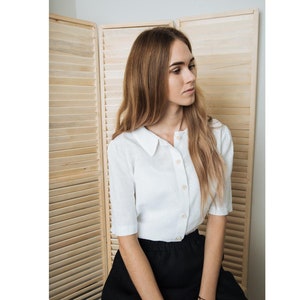 May include: A woman wearing a white linen shirt with a collared neckline and a black skirt. The shirt has a button-up front and short sleeves. The woman is sitting in front of a wooden slat wall.