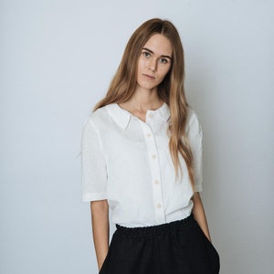 May include: A woman wearing a white collared shirt with a button-up front and black trousers.