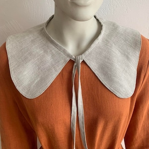 May include: A light gray linen collar with a rounded shape and a tie closure. The collar is worn over a rust-colored linen dress.