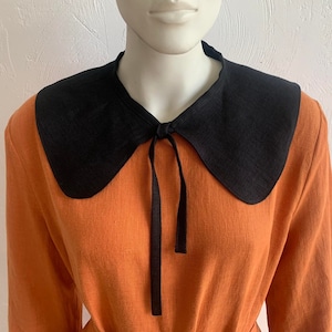 May include: A black linen collar with a rounded shape and a tie at the front. The collar is worn over a burnt orange dress.