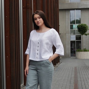 May include: A woman wearing a white linen top with a button-up front and a pair of light blue linen pants. She is wearing white sneakers and has her hand in her pocket.