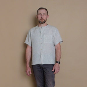 May include: A light grey linen shirt with a mandarin collar and short sleeves. The shirt has a button-up front and a single pocket on the left chest.