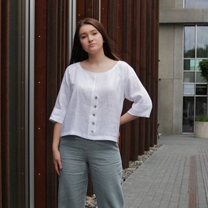 May include: A woman wearing a white linen blouse with a button-up front and a light blue pair of pants.