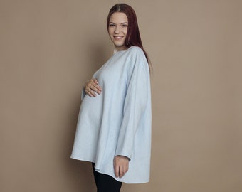 May include: A pregnant person wearing a light blue oversized sweater and black leggings. They are smiling and standing on a light brown background. They are wearing white sneakers.