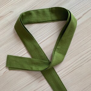 May include: A long, green fabric sash or belt, folded and tied in a knot. The sash is made of a soft, textured material.