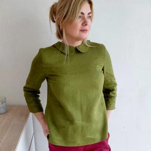 Op de afbeelding: Olijfgroene blouse met lange mouwen en een ronde kraag. De blouse wordt gecombineerd met een bordeauxrode broek. De outfit is eenvoudig en elegant, geschikt voor casual of semi-formele gelegenheden. De blouse heeft een relaxte pasvorm.