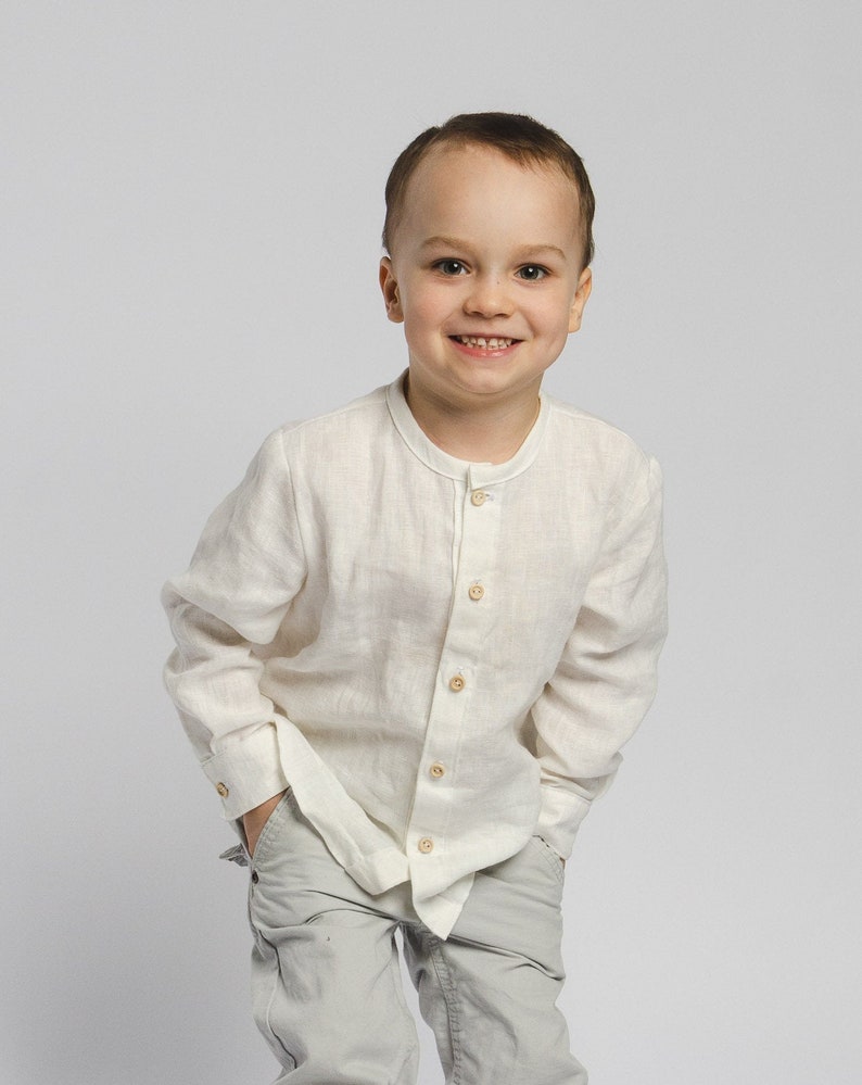 May include: A young boy wearing a white linen shirt with a button-up collar and light gray pants. He is smiling and has his hands in his pockets.
