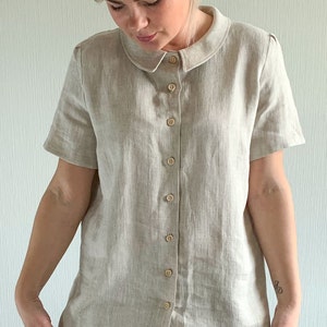 May include: A beige linen shirt with a peter pan collar and short sleeves. The shirt has a button-up front and is made of a lightweight fabric.