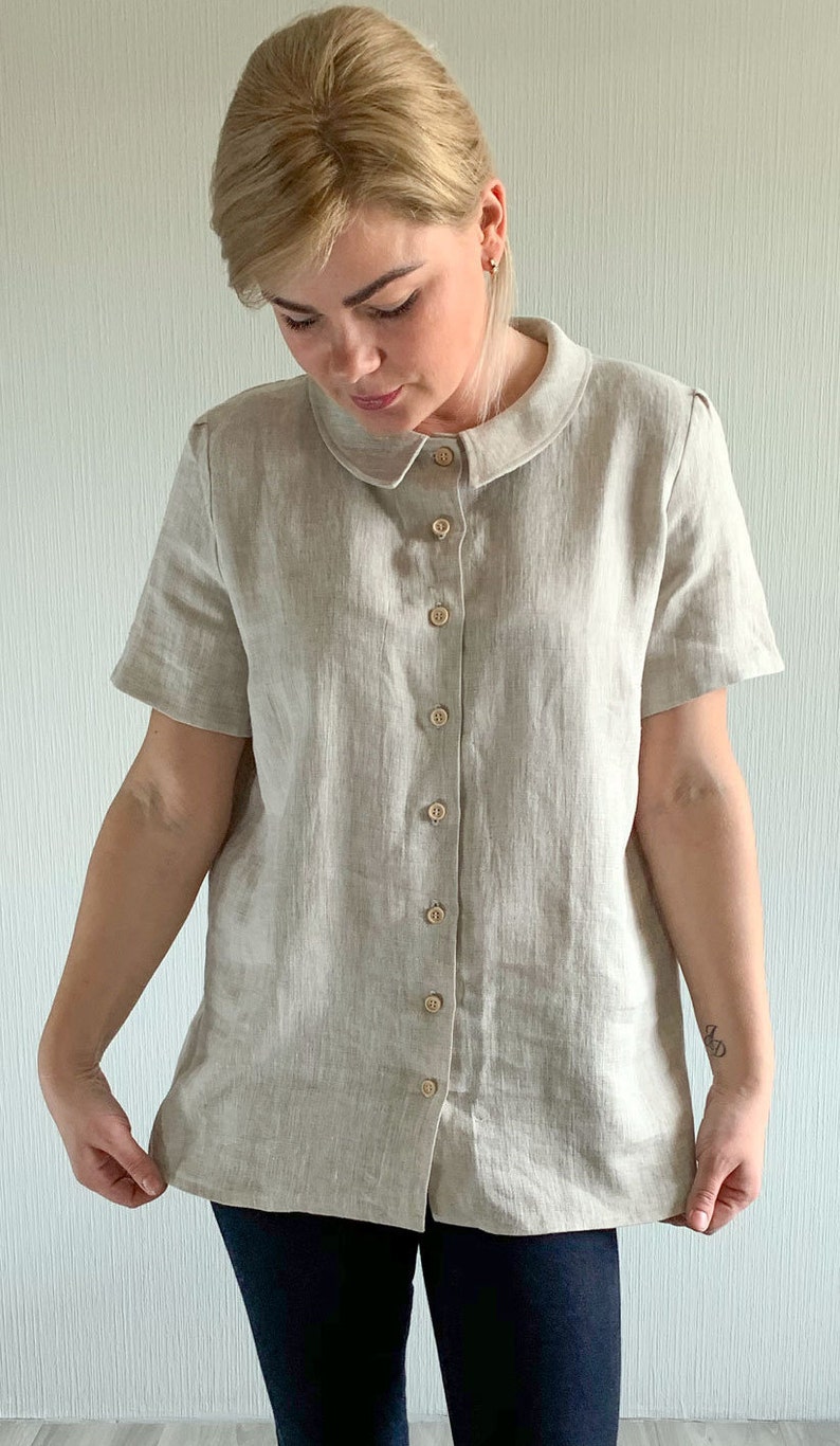 May include: A beige linen shirt with a peter pan collar and short sleeves. The shirt has a button-up front and is made of a lightweight fabric.