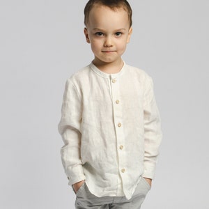May include: A young boy wearing a white linen shirt with a mandarin collar and grey pants. He is standing with his hands in his pockets and looking directly at the camera.